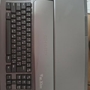 May include: A gray Sharp Font Writer personal word processor with a QWERTY keyboard. The keyboard has various function keys and a numeric keypad. The word processor has the Sharp logo and the words "Font Writer" printed on the front.