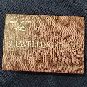 Op de afbeelding: Een vintage "Travelling Chess" set van House Martin. De doos heeft een getextureerd, bruin, reptielachtig patroon met gouden letters. De tekst bevat de merknaam en de productnaam. Een kleine vogelafbeelding staat erboven.
