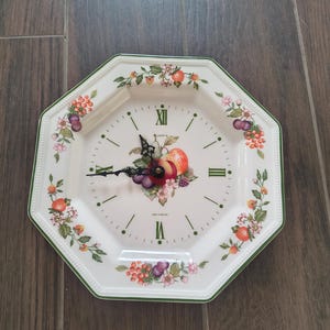 May include: An octagonal wall clock with a cream-colored ceramic face featuring a central fruit illustration, green Roman numerals, and black hands. The border is decorated with a colorful pattern of berries, flowers, and leaves, with the text "MADE IN ENGLAND" printed near the bottom.