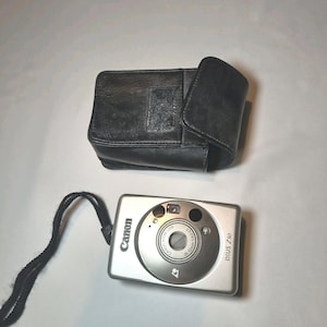 Puede incluir: Cámara digital Canon IXUS Z50 plateada con correa de muñeca negra, junto a una funda de cuero negra. La lente de la cámara tiene un rango de zoom de 26-52 mm.