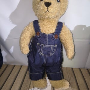 May include: A plush teddy bear wearing denim overalls with brown buttons and white stitching. The bear has a tan fur body, a black nose, and dark eyes. A white tag is attached to the bear's foot.