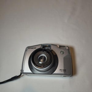 Puede incluir: Cámara digital Polaroid PZ1400 plateada con zoom 1.4x, lente negra y correa de muñeca negra. La cámara tiene flash y objetivo zoom. La cámara está sobre un fondo blanco.