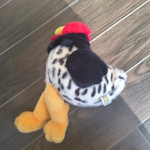May include: A plush rooster toy with a red comb, black wings, and a white and black spotted body. The rooster has yellow feet and a yellow beak. The toy is made by The Manhattan Toy Company.