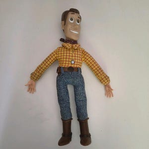 Puede incluir: Una figura de acción de Woody de la franquicia Toy Story. La muñeca tiene una camisa a cuadros amarilla, pantalones vaqueros azules, botas marrones y un cinturón marrón con una hebilla decorativa. La figura tiene un pañuelo rojo y una expresión sonriente.