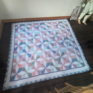 Op de afbeelding: Een handgemaakte quilt met een herhalend patroon van witte, blauwe, groene en koraalkleurige vormen. De quilt heeft een witte rand met een blauw driehoekpatroon. De quilt ligt op een donkerbruine houten vloer.