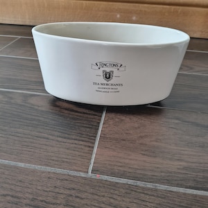 May include: A white, oval ceramic bowl with the text "RINGTONS TEA MERCHANTS" printed on the side. The bowl has a flared design and sits on a dark wood-grain surface. The text includes the address "ALGERNON ROAD NEWCASTLE - TYNE".
