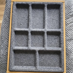 May include: A wooden tray with a gray felt interior divided into nine compartments. The tray has a light brown wooden frame and is designed for organizing small items. The felt provides a soft surface for delicate objects.