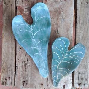 Op de afbeelding: Twee bladvormige decoratieve schalen in verschillende maten, afgewerkt met een matte blauwgroene kleur en voorzien van diep uitgesneden, lichtgekleurde nerven die de natuurlijke structuur van een plantenblad nabootsen op een houten ondergrond.