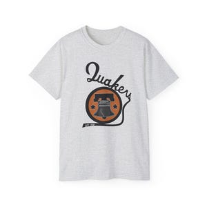 May include: A light gray t-shirt featuring a graphic with the word "Quakers" in a stylized font. The graphic includes a bell inside a circle with stars and the text "Est. 1930".