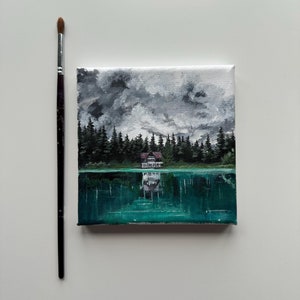 May include: A small painting on canvas depicting a house with a red roof reflected in a lake. The sky is gray and cloudy, and the trees surrounding the lake are dark green.