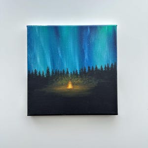 May include: A square canvas painting shows a night forest scene. The sky is a gradient of teal and blue, with streaks of green, suggesting the aurora borealis. Silhouetted trees line the horizon, with a campfire glowing in the centre.