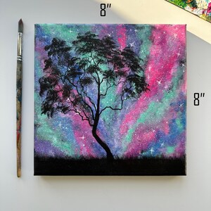 Peut inclure: Une peinture sur toile de 20,3 x 20,3 cm repr&eacute;sentant un arbre en silhouette devant une galaxie color&eacute;e aux teintes roses, violettes et vertes. La galaxie est parsem&eacute;e d'&eacute;toiles blanches.