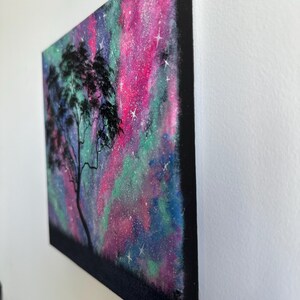 Peut inclure: Une peinture repr&eacute;sentant la silhouette d'un arbre devant une galaxie color&eacute;e aux teintes roses, vertes et bleues. La galaxie est parsem&eacute;e d'&eacute;toiles blanches.