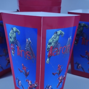 Peut inclure: Boîtes de fête rouge et bleu sur le thème des super-héros. Les boîtes présentent des images de Hulk, Spiderman, Iron Man et Captain America. Le texte sur les boîtes indique "6 Ans" et "Aaron".