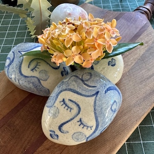 May include: Three ceramic face planters with blue line art on a cream background. The planters hold a bouquet of orange flowers and green leaves. The planters are on a wooden chopping board.