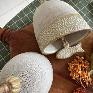 May include: A beige and cream ceramic bell with a textured leaf pattern band and a fish-shaped clapper. The bell has a rope for hanging. The image also shows a cracked ceramic sphere and dried flowers.