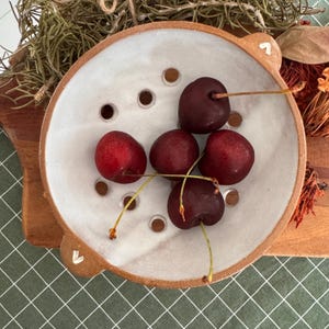 May include: A round ceramic colander with a white interior and a natural terracotta exterior, featuring two small handles with white chevron accents and multiple drainage holes, containing five fresh dark red cherries with stems.