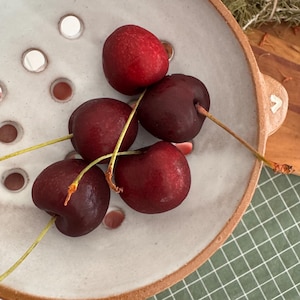 May include: A round ceramic colander with a speckled off-white interior and a natural terracotta rim, featuring evenly spaced drainage holes. Five fresh, dark red cherries with green stems are resting inside the bowl.