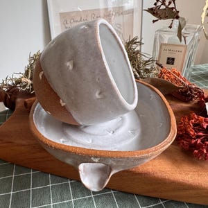 May include: A handmade ceramic cup and saucer set featuring a rustic, unglazed terracotta base and a speckled, off-white glazed upper section. The pieces are decorated with small, raised white heart-shaped accents and have a thick, organic rim.