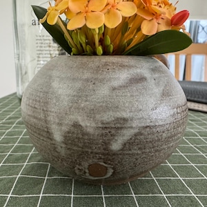 May include: A round, spherical ceramic vase with a textured, dark brown base that transitions into a mottled, light grey glaze toward the top. The vase is filled with a vibrant bouquet of bright orange ixora flowers and green leaves.
