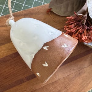 May include: A ceramic bell with a white glazed top and a brown base. The base is decorated with small white heart shapes. A natural twine loop is attached for hanging. The bell is a decorative item.