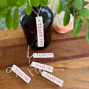 May include: A dark brown glass vase with green leaves and several white rectangular tags. The tags have the words "THANK YOU" printed in red. A seashell is visible in the background.