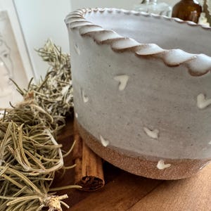 May include: A cylindrical ceramic bowl with a scalloped rim, featuring a light grey upper section decorated with small white heart-shaped indentations and a contrasting unglazed, textured brown base. The bowl has a speckled finish.