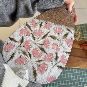 May include: A teardrop-shaped ceramic wall plaque featuring a white textured surface decorated with pink flowers and green leaves. The top section has a brown embossed scroll pattern, and a small hole is present at the top for hanging purposes.