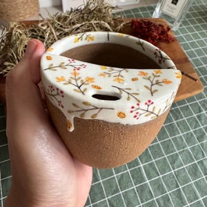 May include: A ceramic mug with a brown, textured clay base and a white glazed upper section. The white rim features a hand-painted pattern of delicate brown branches, small yellow leaves, red berries, and tiny orange ladybugs, with a small slot.