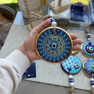 Op de afbeelding: Een ronde, blauw en gouden decoratieve wandhanger met een oogontwerp. De hanger heeft een gouden rand en een touw om op te hangen. Andere soortgelijke items zijn zichtbaar op de achtergrond.