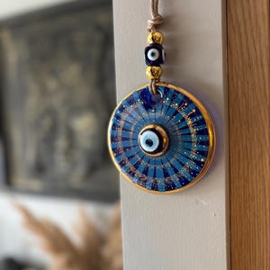 May include: A round, blue and gold decorative charm with an eye design. The charm is suspended by a tan cord with gold beads and a blue cube bead. The charm has a gold border and a detailed blue and gold pattern.
