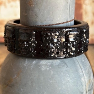 May include: A dark brown, glossy bangle bracelet featuring a repeating, raised relief pattern of Frankenstein's monster faces encircling the entire outer surface. The bracelet has a smooth, polished finish and a thick, circular band construction.