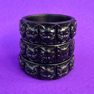 May include: A stack of three black bangle bracelets, each featuring a repeating carved pattern of cat faces around the entire circumference. The bracelets have a smooth, dark finish and are arranged vertically to show the detailed feline design.