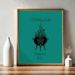 May include: Framed teal artwork with a gold border. The image features a black cauldron with a pentagram, flames, and the text "Witchy stuff happens here" in a decorative font. A white vase and small floral arrangement are in the foreground.