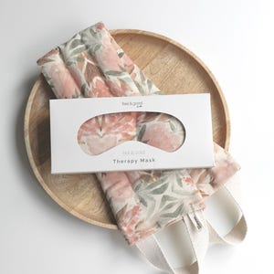 Peut inclure: Un plateau en bois présente une boîte blanche et un masque de thérapie floral. La boîte indique "feel & good co." et "hot & cold Therapy Mask". Le masque et la partie visible de la boîte présentent un motif floral rose, vert et marron. Le masque a des sangles blanches.