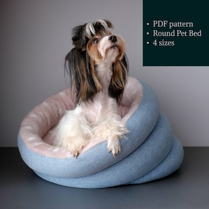 May include: A small dog sits in a blue and pink round pet bed. The bed is made of soft fabric and has a fluffy inner lining. The dog is looking up and has a white, brown, and black coat.