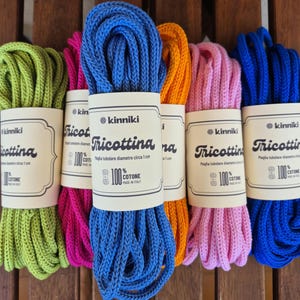 Puede incluir: Ovillos de hilo de algodón de colores verde, rosa, azul y naranja. Cada ovillo está etiquetado con la marca "kinniki" y el texto "Tricottina". El hilo tiene aproximadamente 1 cm de diámetro y está hecho de 100% algodón.