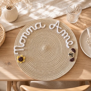 Può includere: Un sottobicchiere rotondo e intrecciato con il nome "Momma Anne" in corsivo bianco. Un fiore all'uncinetto giallo e marrone e un ornamento a forma di orso marrone sono sul sottobicchiere. Il sottobicchiere è su un tavolo di legno.