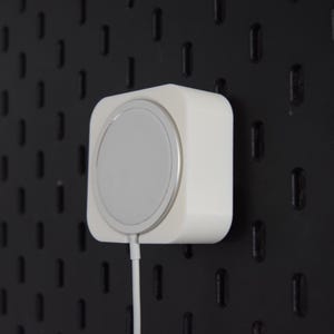 Op de afbeelding: Een witte draadloze oplader met een zilveren oplaadoppervlak is gemonteerd op een zwart pegboard. Een witte kabel loopt van de oplader. De oplader heeft een vierkant lichaam met afgeronde randen.