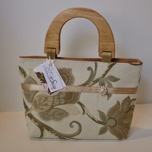 K&ouml;nnte beinhalten: Eine beige Handtasche mit Blumenmuster und einem Holzgriff. Die Tasche hat ein braunes Band mit einem dekorativen Knopf. Das Etikett auf der Tasche lautet "Sassy & Handmade".