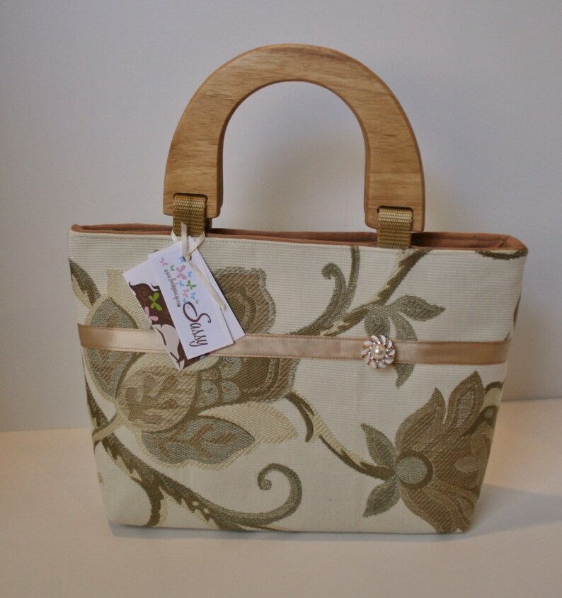 K&ouml;nnte beinhalten: Eine beige Handtasche mit Blumenmuster und einem Holzgriff. Die Tasche hat ein braunes Band mit einem dekorativen Knopf. Das Etikett auf der Tasche lautet "Sassy & Handmade".