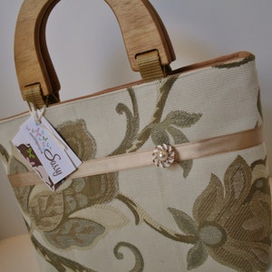 K&ouml;nnte beinhalten: Eine beige Handtasche mit Blumenmuster und einem Holzgriff. Die Tasche hat ein beigefarbenes Band mit einem perlmuttartigen Knopf. Die Tasche hat ein Etikett mit der Aufschrift "Sassy".
