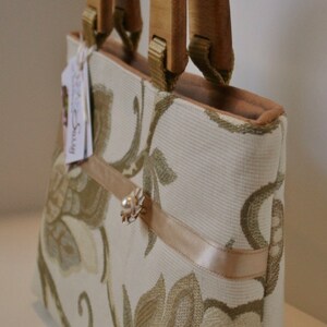 K&ouml;nnte beinhalten: Eine beige Handtasche mit Blumenmuster und einem Holzgriff. Die Tasche hat ein beigefarbenes Satinband und eine kleine Perlenverzierung.