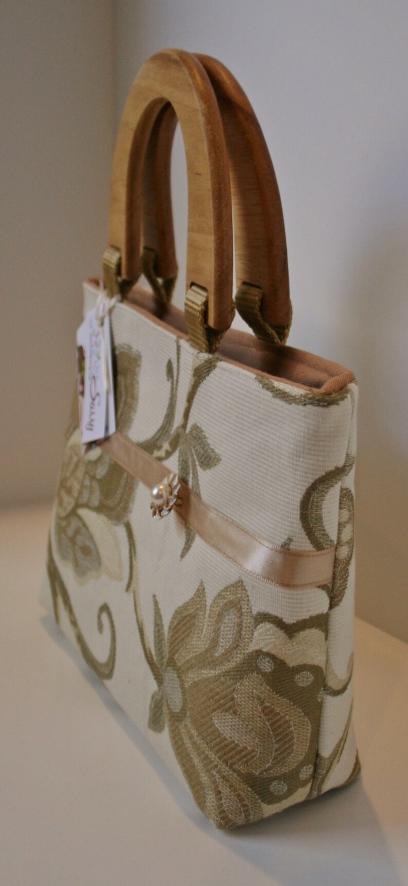 K&ouml;nnte beinhalten: Eine beige Handtasche mit Blumenmuster und einem Holzgriff. Die Tasche hat ein beigefarbenes Satinband und eine kleine Perlenverzierung.