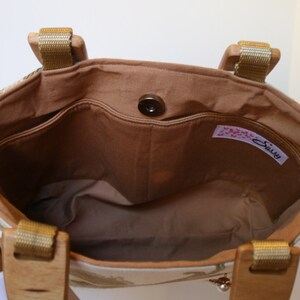 K&ouml;nnte beinhalten: Das Innere einer beige Stofftasche mit braunem Futter. Die Tasche hat einen braunen Knopfverschluss und ein Etikett mit der Aufschrift "Sally".