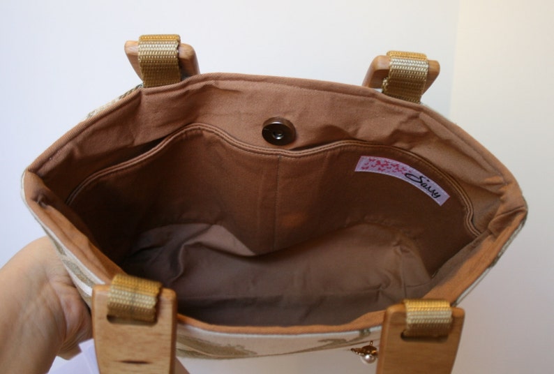 K&ouml;nnte beinhalten: Das Innere einer beige Stofftasche mit braunem Futter. Die Tasche hat einen braunen Knopfverschluss und ein Etikett mit der Aufschrift "Sally".