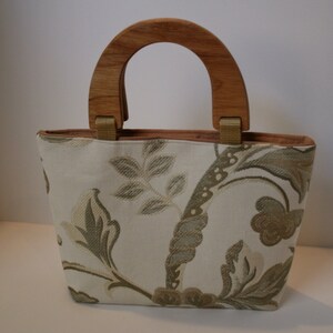 K&ouml;nnte beinhalten: Eine wei&szlig;e Handtasche mit Blumenmuster und einem Holzgriff. Die Tasche hat ein braunes Futter.