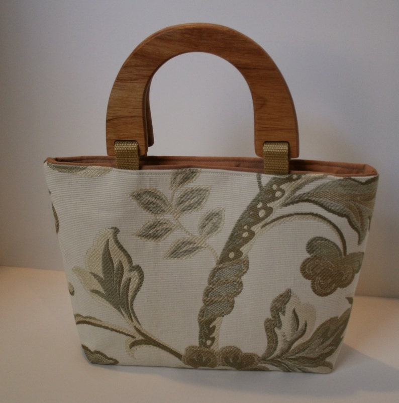 K&ouml;nnte beinhalten: Eine wei&szlig;e Handtasche mit Blumenmuster und einem Holzgriff. Die Tasche hat ein braunes Futter.