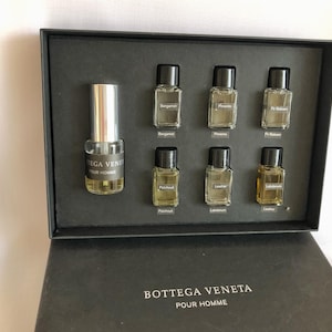 May include: A Bottega Veneta Pour Homme fragrance set in a black box. The set includes a full-size bottle and several smaller sample bottles with labels indicating various scents like Bergamot, Patchouli, and Leather. The box is open, revealing the contents.