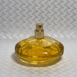 Könnte beinhalten: Ein klarer Glasparfümflakon mit einem goldfarbenen Zerstäuber. Die Flasche ist mit einer gelben Flüssigkeit gefüllt. Die Flasche hat eine abgerundete, ovale Form. Der Hintergrund ist eine weiße, strukturierte Oberfläche.